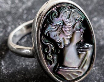 Könnte beinhalten: Ein silberner Ring mit einem ovalen schwarzen und irisierenden Perlmutt-Kamee, der das Profil einer Frau zeigt.