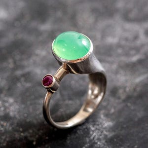 Peut inclure: Une bague en argent avec une pierre précieuse verte et une plus petite pierre rouge. La bague a un design unique avec la plus grande pierre sertie dans une bélière surélevée.