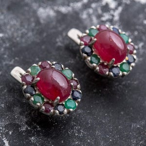 May include: A pair of silver earrings with red ruby gemstones surrounded by smaller green and blue gemstones.