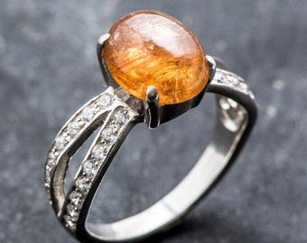 May include: A silver ring featuring a large, oval-cut, orange gemstone. The band splits into two parallel paths, each lined with small, clear gemstones. The ring is set against a dark gray background.