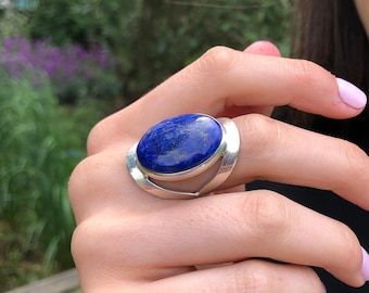 May include: A silver ring with a large oval lapis lazuli gemstone set in a silver band. The ring has a unique design with a curved band that wraps around the stone.