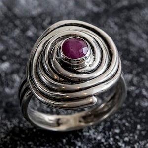 Peut inclure: Bague en argent avec un motif en spirale et une pierre précieuse rouge foncé ronde au centre. La monture de la bague présente un aspect texturé et superposé, la pierre est sertie dans une lunette simple.