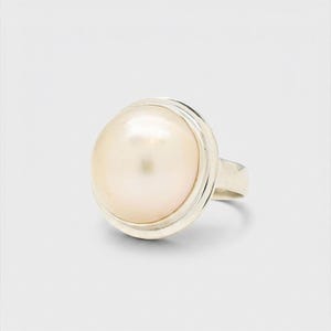 May include: A ring featuring a large, round, cream-colored pearl set in a polished silver-toned metal band. The pearl has a smooth, lustrous surface and is securely held within a simple, circular bezel setting that highlights its natural shape.