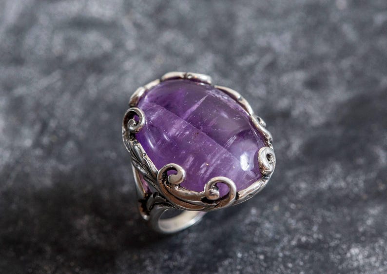 May include: A silver ring with an oval-shaped amethyst gemstone set in a decorative filigree design.