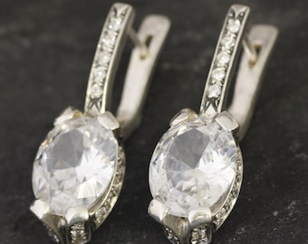 May include: A pair of silver earrings featuring a clear round stone set in a silver setting. The earrings have a simple design and are accented with small clear stones.