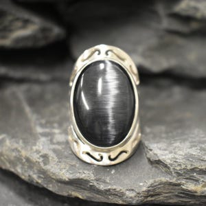 May include: A silver ring with a large oval black gemstone. The ring has a decorative design around the stone.