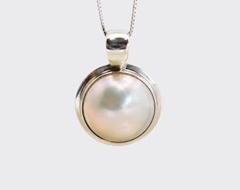 Puede incluir: Un colgante de perla blanca iridiscente de forma redonda, engastado en un bisel de metal pulido de tono plateado. El colgante cuelga de una delicada cadena plateada y presenta una superficie lisa y brillante que refleja la luz con tonos rosados y azulados.