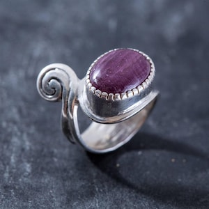 May include: A silver ring with a purple gemstone set in a bezel setting. The ring has a unique, swirling design.