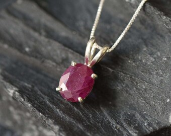 May include: A pendant necklace featuring an oval-cut, deep red gemstone held in a four-prong silver setting. The pendant is attached to a delicate silver box chain, showcasing a faceted surface that reflects light across the stone's dark red hue.