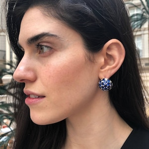 May include: A woman wearing a pair of blue gemstone earrings. The earrings are round and have a cluster of blue gemstones set in a silver metal setting.