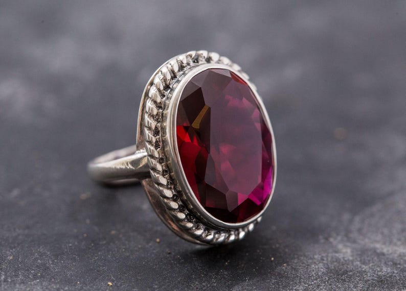 May include: A silver ring with a large, oval-shaped, deep red gemstone set in a decorative, rope-like border.
