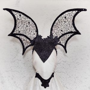 May include: A black lace bat wing headpiece with a matching bodice and collar. The wings are detailed with a delicate web-like pattern. The headpiece is displayed on a white mannequin head, showcasing the gothic and fantasy aesthetic.