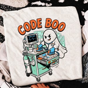 May include: A white t-shirt featuring a graphic of a ghost dressed as a medical professional using a defibrillator on a medical cart. The text CODE BOO is printed in orange above the ghost. The medical equipment is depicted in shades of green and blue.