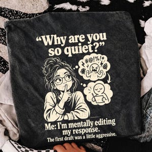 Op de afbeelding: Een donkergrijs t-shirt met een afbeelding van een persoon met een bril en een knotje die een vinger voor de lippen houdt. De tekst luidt: Why are you so quiet? Me: I'm mentally editing my response. The first draft was a little aggressive.