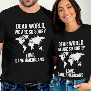 以下が含まれることがあります： 白いグラフィックデザインが施された黒いTシャツ2枚。グラフィックには世界地図と「Dear World, We Are So Sorry Love, Sane Americans」というテキストが描かれています。