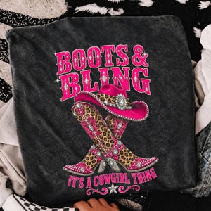 May include: A charcoal gray t-shirt featuring a graphic print of a pink cowboy hat and two crossed cowboy boots with a leopard print pattern. The design includes the text "BOOTS & BLING" and "IT'S A COWGIRL THING" in bright pink lettering.
