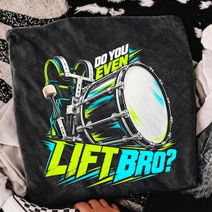 May include: A charcoal gray t-shirt featuring a graphic of a marching bass drum with a harness and mallet. The text reads DO YOU EVEN LIFT BRO? in bright lime green and cyan blue lettering with motion lines surrounding the drum illustration.