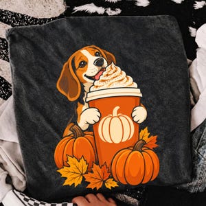 Puede incluir: Una camiseta gris carbón oscuro con una ilustración gráfica de un perro beagle lamiendo nata montada de un vaso naranja alto. El vaso está decorado con un icono de calabaza blanca, y el diseño incluye dos calabazas naranjas y hojas de otoño.