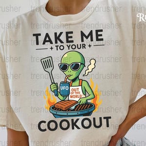 Puede incluir: Camiseta color crema con el texto "TAKE ME TO YOUR COOKOUT". El diseño presenta un alienígena verde con gafas de sol, asando comida y bebiendo de una lata "UFO". El alienígena lleva un delantal que dice "OUT OF THIS WORLD".