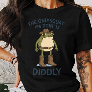 Pode incluir: Camiseta preta com um sapo de desenho animado vestindo um chapéu de cowboy e botas. O texto "The only squat I'm doin' is diddly" está impresso na camiseta.