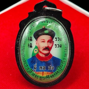 May include: A black pendant with a portrait of a man in a blue and red uniform, a black hat, and a mustache. The portrait is set against a green background with text. The pendant has a black frame and a silver loop.