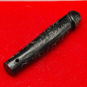 Può includere: Un oggetto cilindrico nero con disegni incisi, forse un timbro o un elemento decorativo, poggia su una superficie di velluto rosso all'interno di un contenitore rettangolare bianco. L'oggetto ha un piccolo foro a un'estremità.