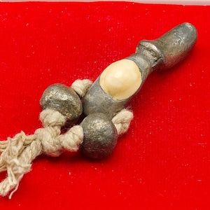 Può includere: Un oggetto metallico color argento con un intarsio color crema e due sfere attaccate, collegate da una corda beige annodata. L'oggetto poggia su una superficie di velluto rosso all'interno di una custodia di plastica bianca, su sfondo nero.