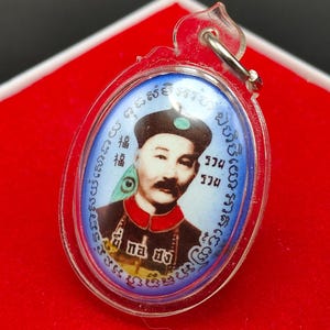 May include: A round, clear plastic pendant with a blue and white image of a man in a traditional Chinese hat. The image is surrounded by a blue border with Thai text. The pendant has a silver loop at the top.