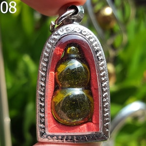 Puede incluir: Un colgante de plata con un amuleto de calabaza de vidrio verde en su interior. El colgante está en un marco de plata con un fondo rojo.