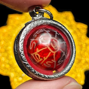 May include: A round, silver pendant with a red center and etched gold lettering. The pendant is held between two fingers against a blurred yellow background. The pendant has a silver loop at the top.