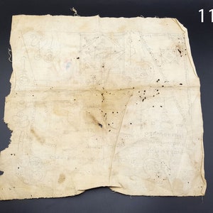 May include: A faded, off-white cloth with a handwritten design in black ink. The cloth is folded in the middle and has several small black spots. The cloth measures 11 inches in length.