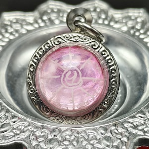 May include: A close-up of a silver pendant with a pink, circular center. The center has a white, etched design. The pendant is set in a decorative silver frame and has a small loop at the top for a chain.