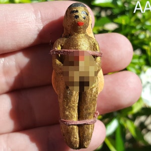Puede incluir: Una pequeña figurita dorada de una persona con boca roja y ojos negros. La figurita está atada con una cuerda marrón alrededor de la cintura y el pecho. La figurita sostiene un objeto redondo frente a su cuerpo.