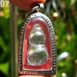 Puede incluir: Un colgante de plata con un amuleto de calabaza de vidrio rojo y transparente. El colgante tiene la forma de un amuleto tradicional tailandés.