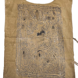 Op de afbeelding: Tan-kleurig stoffen kledingstuk met een ronde hals en ingewikkelde zwarte inkt illustraties. Het kunstwerk toont figuren, dieren en script, waardoor een gedetailleerd en cultureel ontwerp ontstaat. Het kledingstuk heeft touwtjes aan de zijkanten.