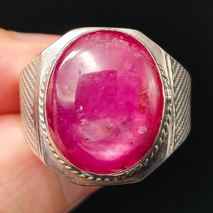 Op de afbeelding: Een zilveren ring met een grote, ovaalvormige, in cabochon geslepen magentakleurige edelsteen, gezet in een rand met een gedraaid touwpatroon. De ringband heeft aan de zijkanten een textuur met een kruisarcering voor een gedetailleerde afwerking.