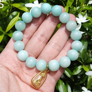 Puede incluir: Un brazalete de piedras preciosas azul claro con un dije de oro. El brazalete está hecho de cuentas redondas y tiene un colgante con una pequeña figura dorada.