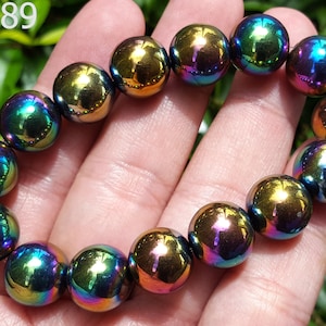 Könnte beinhalten: Ein Armband aus irisierenden, kugelförmigen Perlen. Die Perlen reflektieren ein Farbspektrum, darunter Gold, Grün, Blau und Lila. Das Armband wird vor einem Hintergrund aus grünem Laub gezeigt. Der Text "AA2889" befindet sich in der oberen linken Ecke.