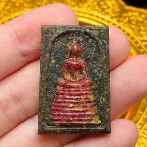May include: A small, rectangular amulet featuring a seated Buddha figure. The Buddha is depicted in red and gold tones, with a detailed layered robe. The amulet has a dark, textured background and a decorative border. The overall aesthetic suggests a religious or spiritual item.