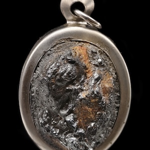 May include: Oval-shaped pendant with a silver-colored frame and a textured, dark gray center. The center has a rough, uneven surface with hints of brown and orange. A small loop is at the top for a chain.