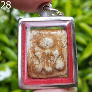 Puede incluir: Un colgante de plata con un borde rojo y un frente de vidrio transparente. El colgante contiene un pequeño amuleto rectangular blanco y marrón.