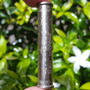 May include: A silver metal tube with intricate engravings. The tube is divided into two sections and appears to be a charm or amulet.