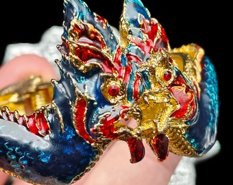 Könnte beinhalten: Ein goldfarbenes Armband mit einem detaillierten Design aus zwei mythischen Kreaturen mit roter, blauer und goldener Emaille. Die Kreaturen haben komplizierte Details und eine strukturierte Oberfläche.