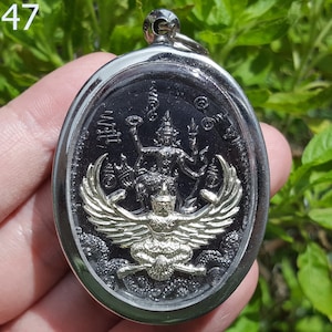 May include: A silver oval pendant with a black and gold image of a mythical creature with wings. The creature is sitting on a cloud and has a crown on its head. The pendant has Thai writing around the edge.
