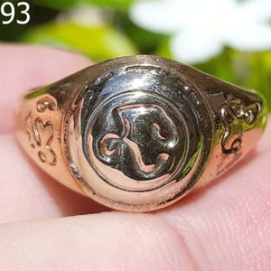 Op de afbeelding: Een gouden ring met een ingewikkeld ontwerp met een symbool in het midden. De ring wordt aan een vinger gedragen.