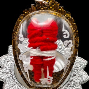 Puede incluir: Un amuleto rojo tejido, encerrado en un colgante transparente de forma ovalada. El amuleto está envuelto con hilo blanco y colocado sobre una base plateada ornamentada. El colgante tiene un borde dorado y está suspendido de un pequeño lazo.