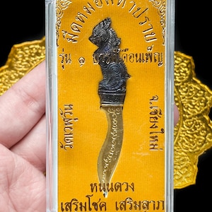 May include: A small, ornate knife encased in a clear plastic case. The knife has a curved blade and a dark handle with intricate carvings. The background is a gold-colored card with Thai script.