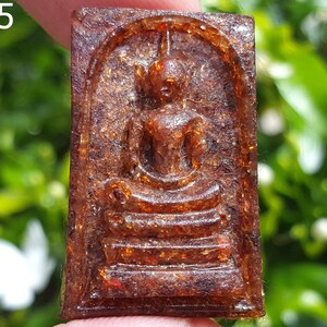 May include: A small, rectangular, brown amulet with a raised image of a seated Buddha. The amulet is made of a translucent material with a speckled appearance.