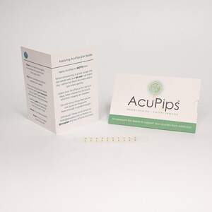 May include: A close-up of a white card with instructions on how to apply ear seeds for acupressure. The card is next to a small white package with the text "AcuPips Modern Healing Ancient Practice Acupressure Ear Seeds to support your journey from addiction". The package contains a strip of 10 small gold ear seeds.