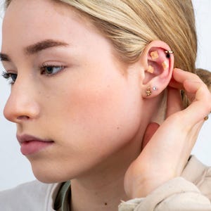 May include: A close-up of a person's ear with multiple gold earrings. The earrings include a small star, a small circle, and a larger hoop with a small black bead. There are also two small square-shaped adhesive bandages on the earlobe.